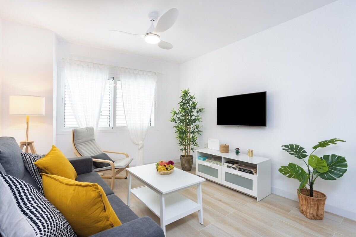 Ein modernes Wohnzimmer mit einem grauen Sofa, gelben Kissen, einem weißen Couchtisch, einer weißen TV-Konsole und einer großen Pflanze.