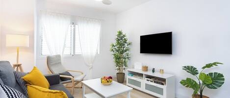 Ein modernes Wohnzimmer mit einem grauen Sofa, gelben Kissen, einem weißen Couchtisch, einer weißen TV-Konsole und einer großen Pflanze.
