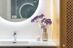 Ein Badezimmer mit einem weißen Waschbecken, einem runden Spiegel und einer Vase mit violetten Blumen.