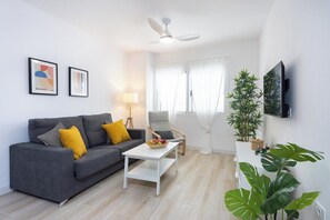 Ein modernes Wohnzimmer mit grauem Sofa, weißem Couchtisch, Stehlampe, Deckenventilator und einem an der Wand montierten Fernseher.