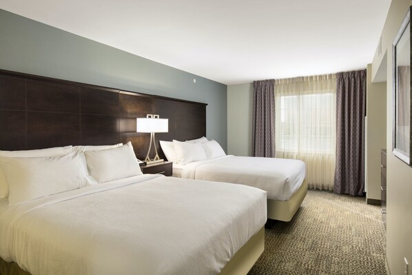 Habitación de hotel con dos camas, una mesita de noche con lámpara, ventana con cortinas y suelo alfombrado.