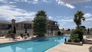 A swimming pool with lounge chairs and umbrellas, surrounded by a fence and palm trees. - Darling 2 Bed 2 Bath Condo in Sports Village - Updated! (St. George)