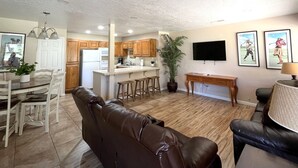 A modern living room with a brown leather sofa, a dining area with white chairs, a kitchen with wooden cabinets, and a TV mounted on the wall. - Darling 2 Bed 2 Bath Condo in Sports Village - Updated! (St. George)