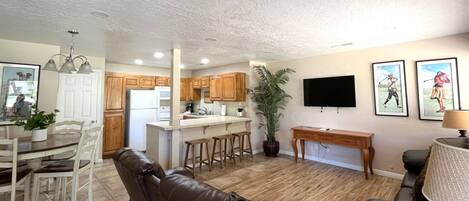 A modern living room with a brown leather sofa, a dining area with white chairs, a kitchen with wooden cabinets, and a TV mounted on the wall.