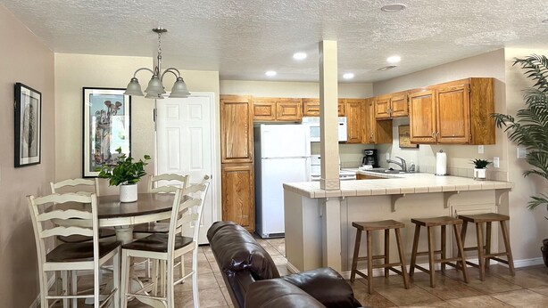 A modern kitchen with wooden cabinets, a central island, and a dining area with a bench and chairs. - Darling 2 Bed 2 Bath Condo in Sports Village - Updated! (St. George)