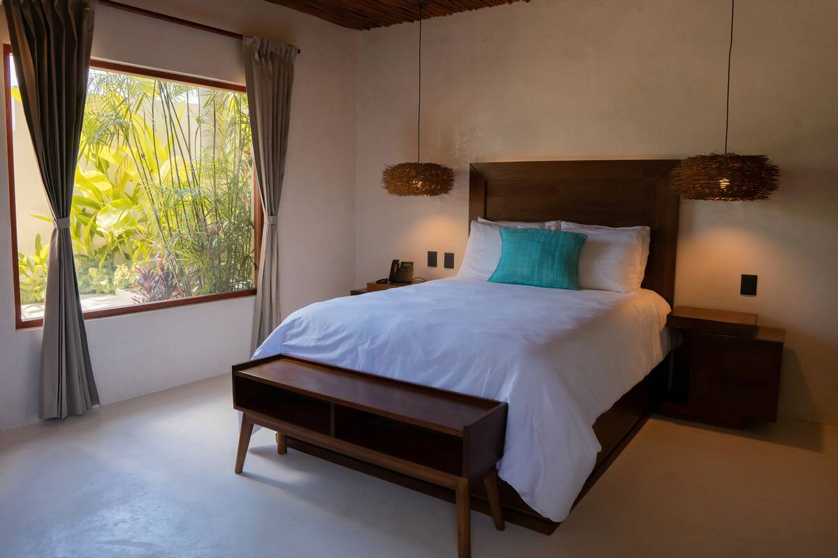 Een slaapkamer met een groot bed, een houten hoofdeinde, nachtkastjes, een bankje en uitzicht op groen door het raam.