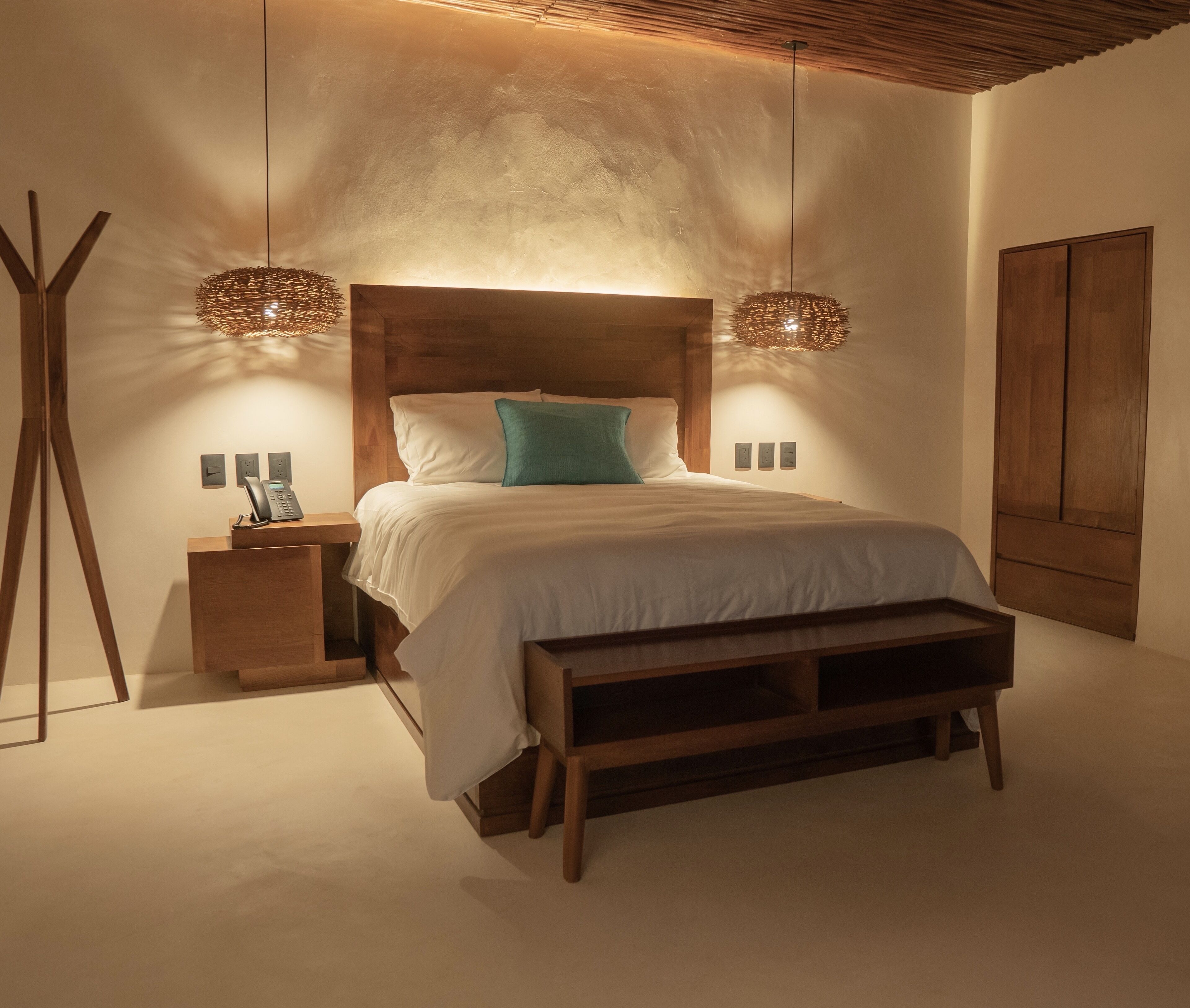 Un dormitorio moderno con cabecera de madera, mesita de noche con teléfono, banco al pie de la cama y dos lámparas colgantes.