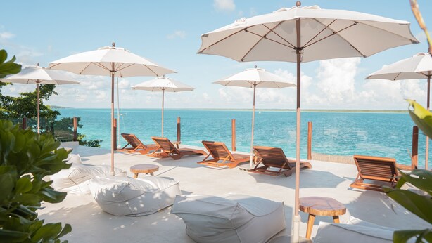 A beachside lounge area with white umbrellas, wooden lounge chairs, and white cushions.