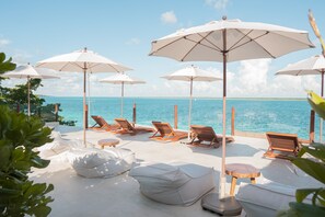 Un área de descanso en la playa con sombrillas blancas, sillones de madera y cojines blancos.