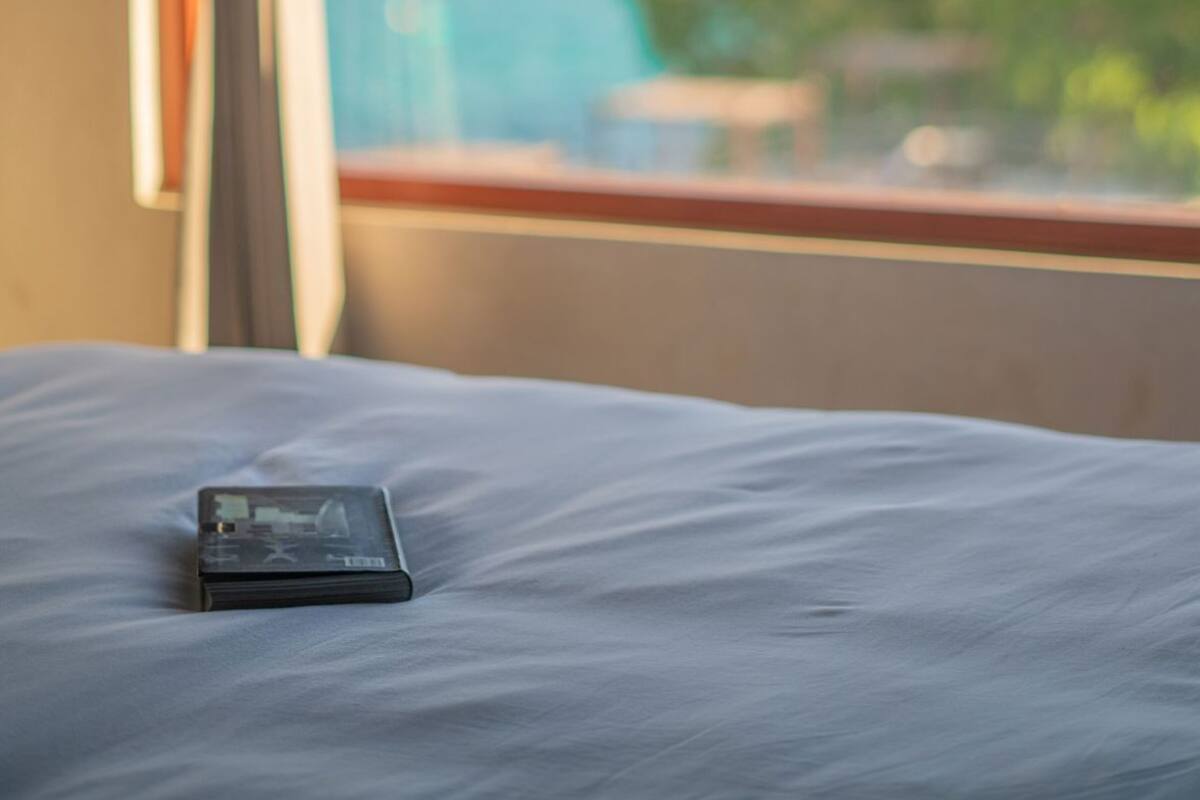 Een bed met een smartphone erop, een raam met gordijnen, en uitzicht op de oceaan en groen.