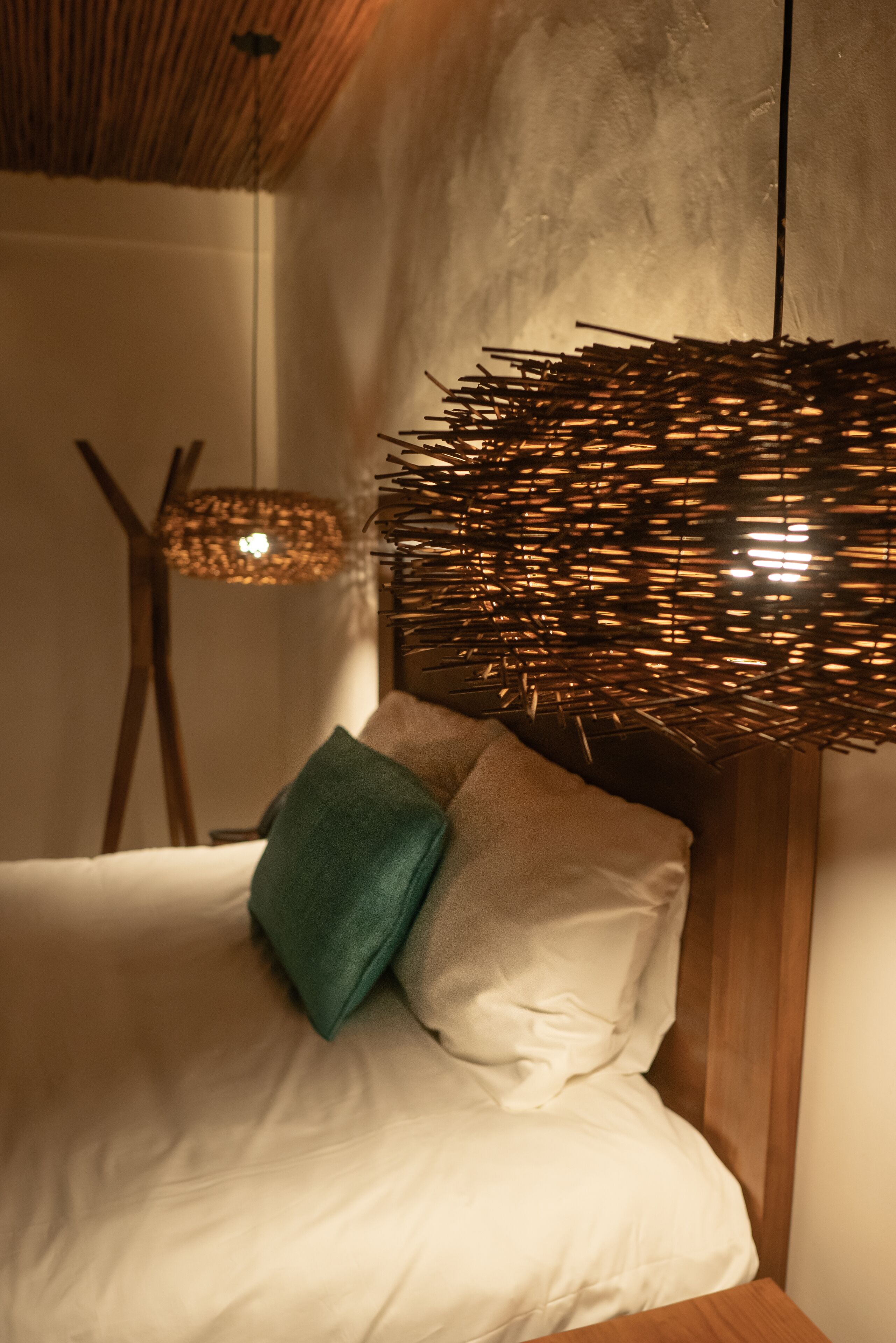Un lit avec une literie blanche et un oreiller vert, une lampe en osier et une tête de lit en bois.