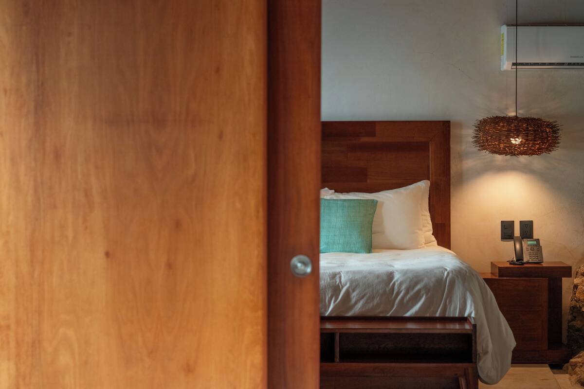 Een slaapkamer met een houten deur, een bed met witte lakens, een nachtkastje met een lamp, en een wandairconditioner.