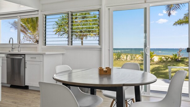 A modern kitchen with white cabinets, a stainless steel dishwasher, and a round dining table with white chairs. Large windows offer a view of the ocean and palm trees.