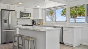 A modern kitchen with white cabinetry, stainless steel appliances, a large island, and a view of palm trees through a window.