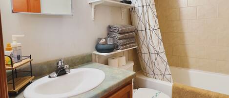 Une salle de bain avec un double lavabo, des toilettes, une douche avec un rideau à motifs et une étagère contenant des articles de toilette.