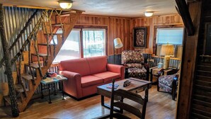 A cozy cabin interior with a wooden staircase, a red sofa, a patterned armchair, and a dining table.