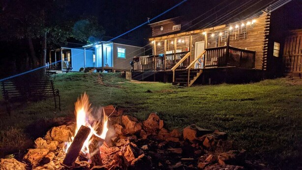 A cabin with a fire pit in the foreground, a porch with lights, and a small building in the background.