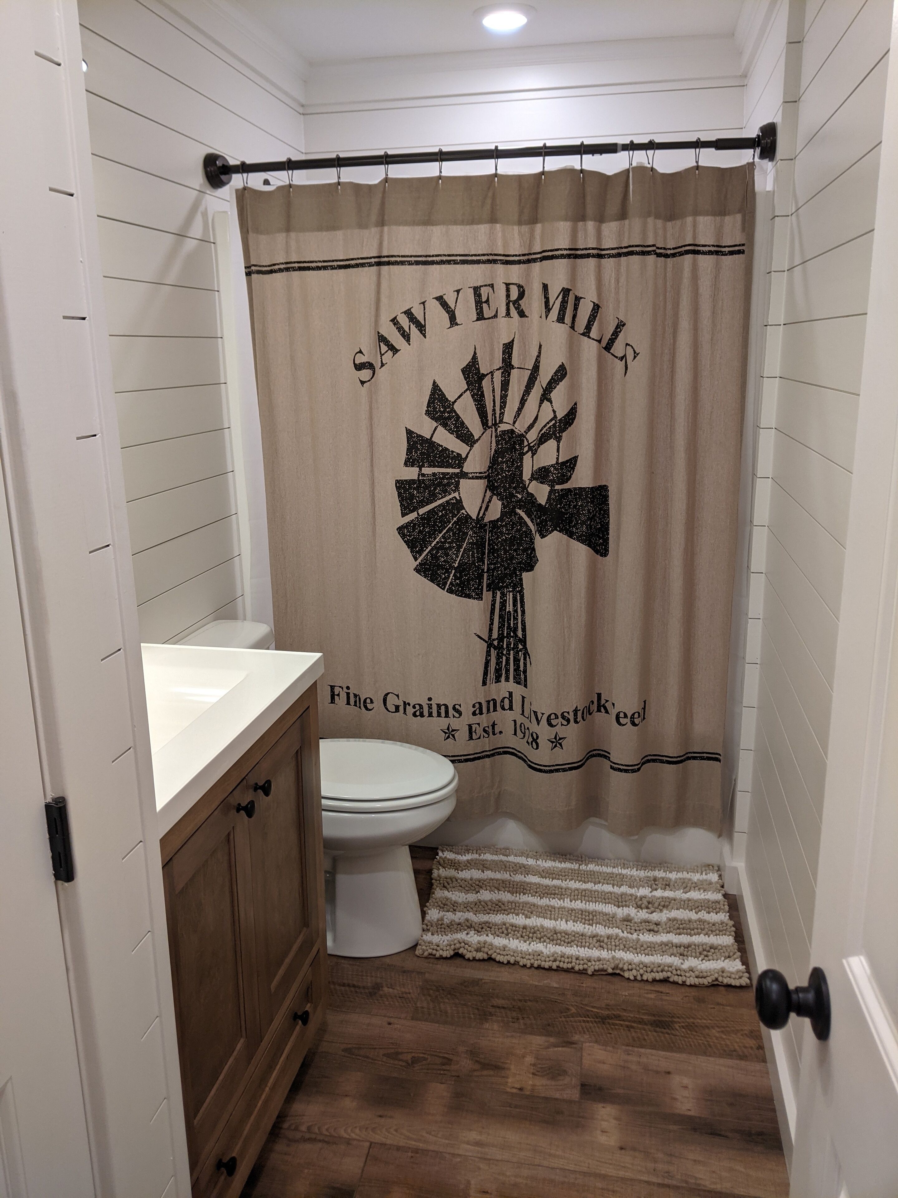 Een badkamer met douchegordijn met molen dessin en tekst, een toilet, houten kastjes en een witte wastafel.
