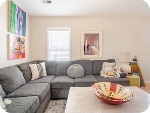 A living room with a grey sectional sofa, a coffee table, and a colorful bowl. There are two paintings on the wall and a window with blinds. - Cozy Beach Cottage DM for winter prices king beds (Oak Island)