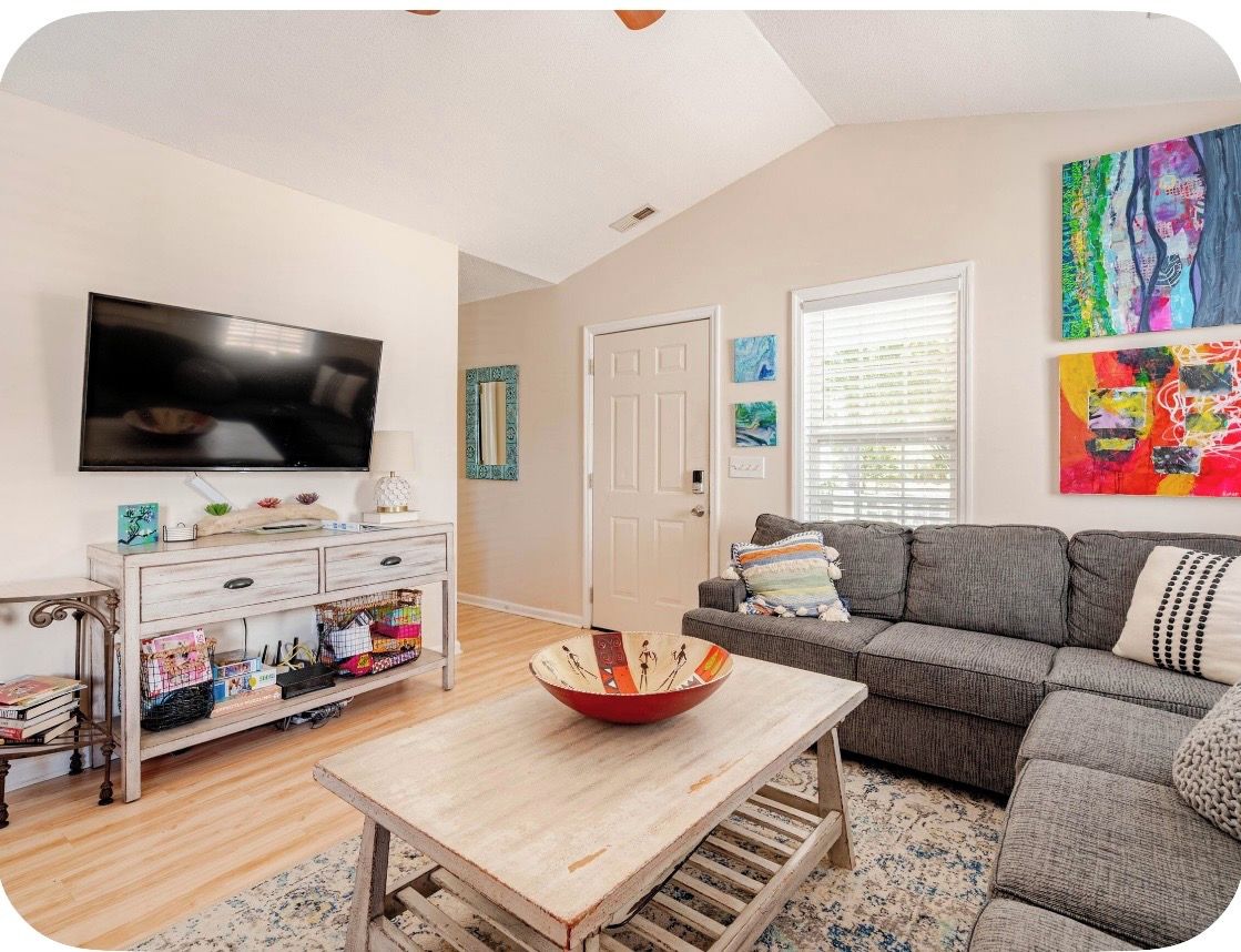 A living room with a flat-screen TV, a wooden coffee table, a gray sofa, and colorful artwork on the walls.