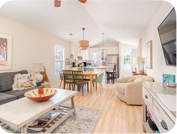 A modern living room with a sofa, dining table, and chairs, a kitchen area with cabinets and appliances, and a TV mounted on the wall.