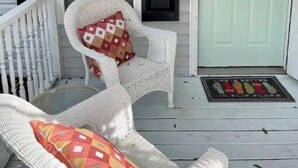 A porch with two wicker chairs, one with a patterned cushion and another with a solid cushion. A white door with a green mat is visible.