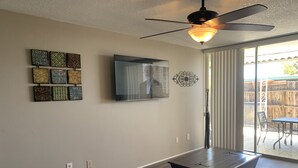 Una sala de estar con ventilador de techo, un televisor de pantalla plana montado en la pared y una mesa de comedor con sillas.