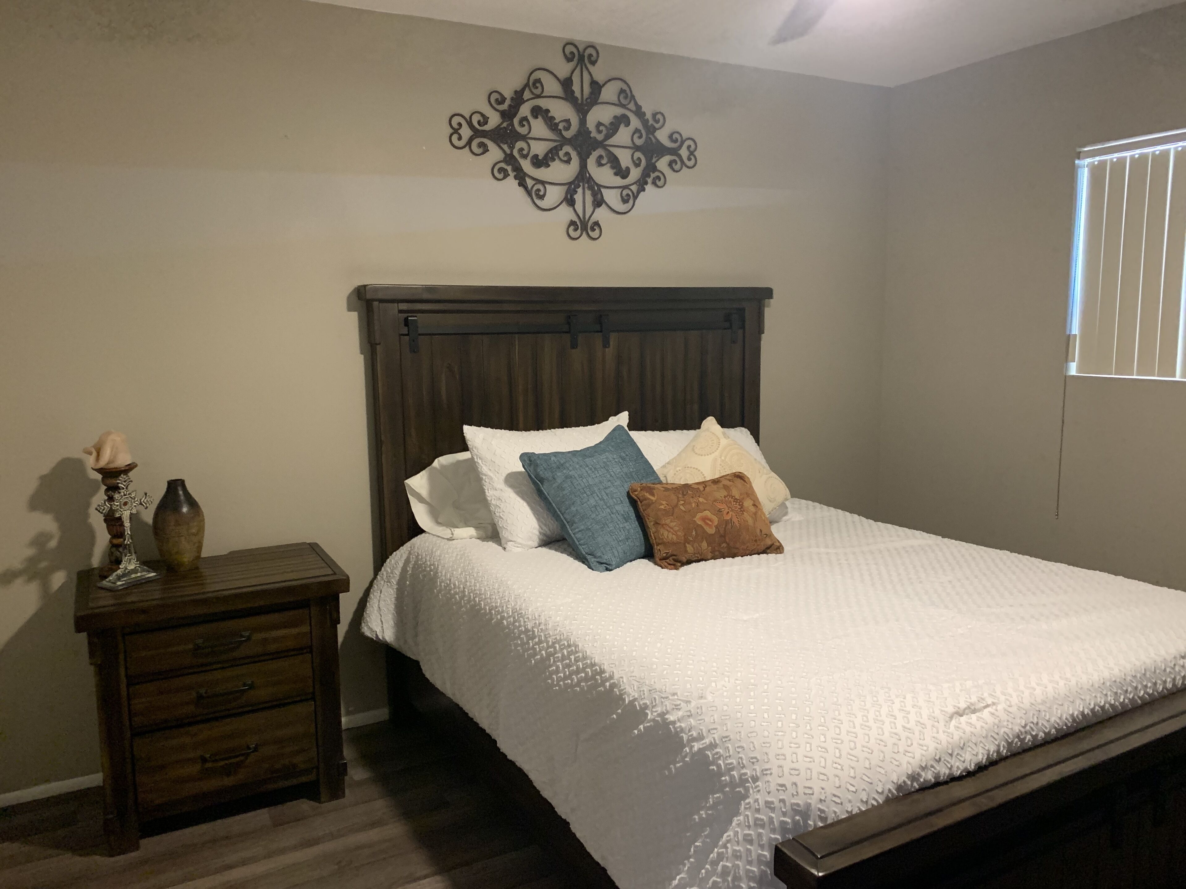 A bedroom with a wooden bed, white bedding, a nightstand with a vase, and a decorative wall piece.