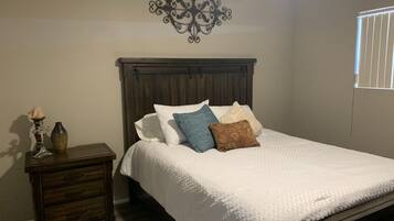 A bedroom with a wooden bed, white bedding, a nightstand with a vase, and a decorative wall piece.