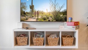 Una televisione fissata al muro mostra una scena desertica con cactus e due persone che camminano. Sotto la TV, ci sono cesti portaoggetti e una scatola con l'etichetta "Catan".