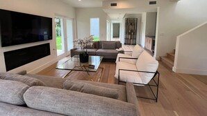 Ein modernes Wohnzimmer mit einem Flachbildfernseher, grauen Sofas, einem Glastisch und einer Treppe, die zu einer weiteren Ebene führt.