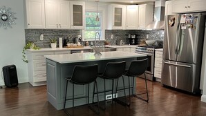 A modern kitchen with white cabinets, a stainless steel refrigerator, a large island with bar stools, and a wooden floor.