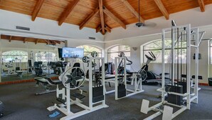 Academia com diversos equipamentos de exercício, espelhos grandes e uma televisão.