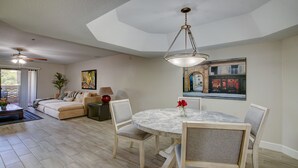 Uma sala de estar ampla com um sofá grande, uma mesa de jantar com cadeiras, um ventilador de teto e um quadro de uma cena urbana.