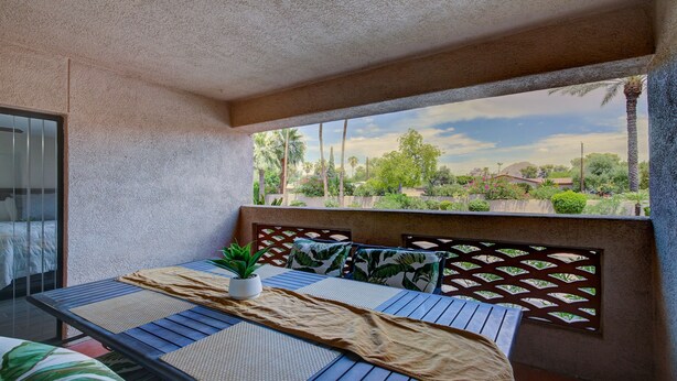 Varanda com vista para um jardim paisagístico, uma mesa com uma planta e uma cama com cabeceira estampada.