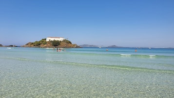 Uma praia com águas cristalinas, um edifício branco numa pequena ilha e algumas pessoas na água.