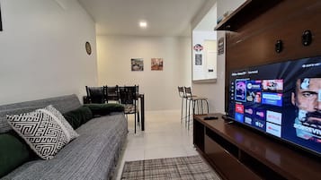 Uma sala de estar com sofá, cadeiras, televisão e prateleira na parede.