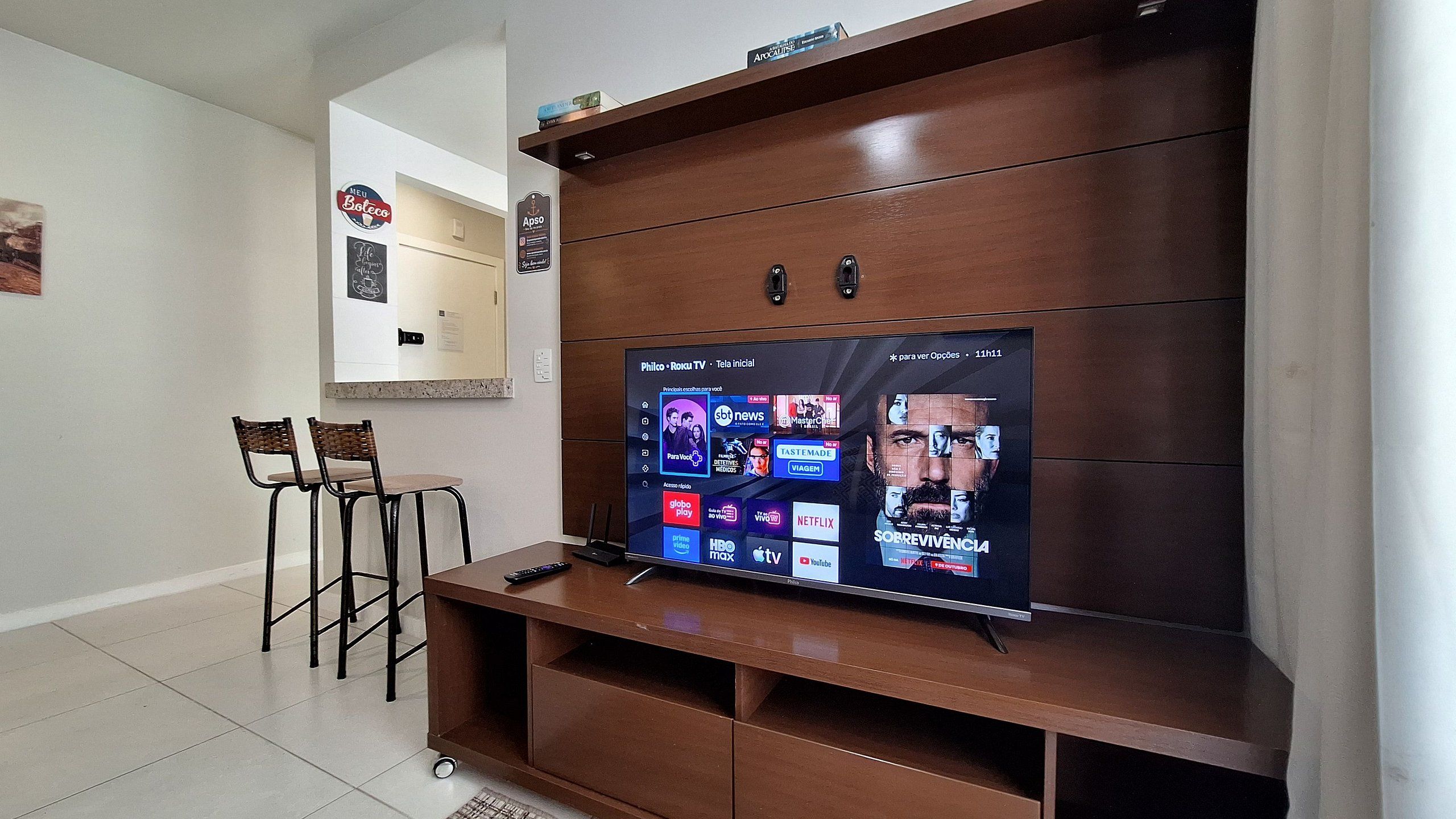 A living room with a flat-screen TV mounted on a wooden cabinet, a coffee table, two chairs, and a small table with a remote control.