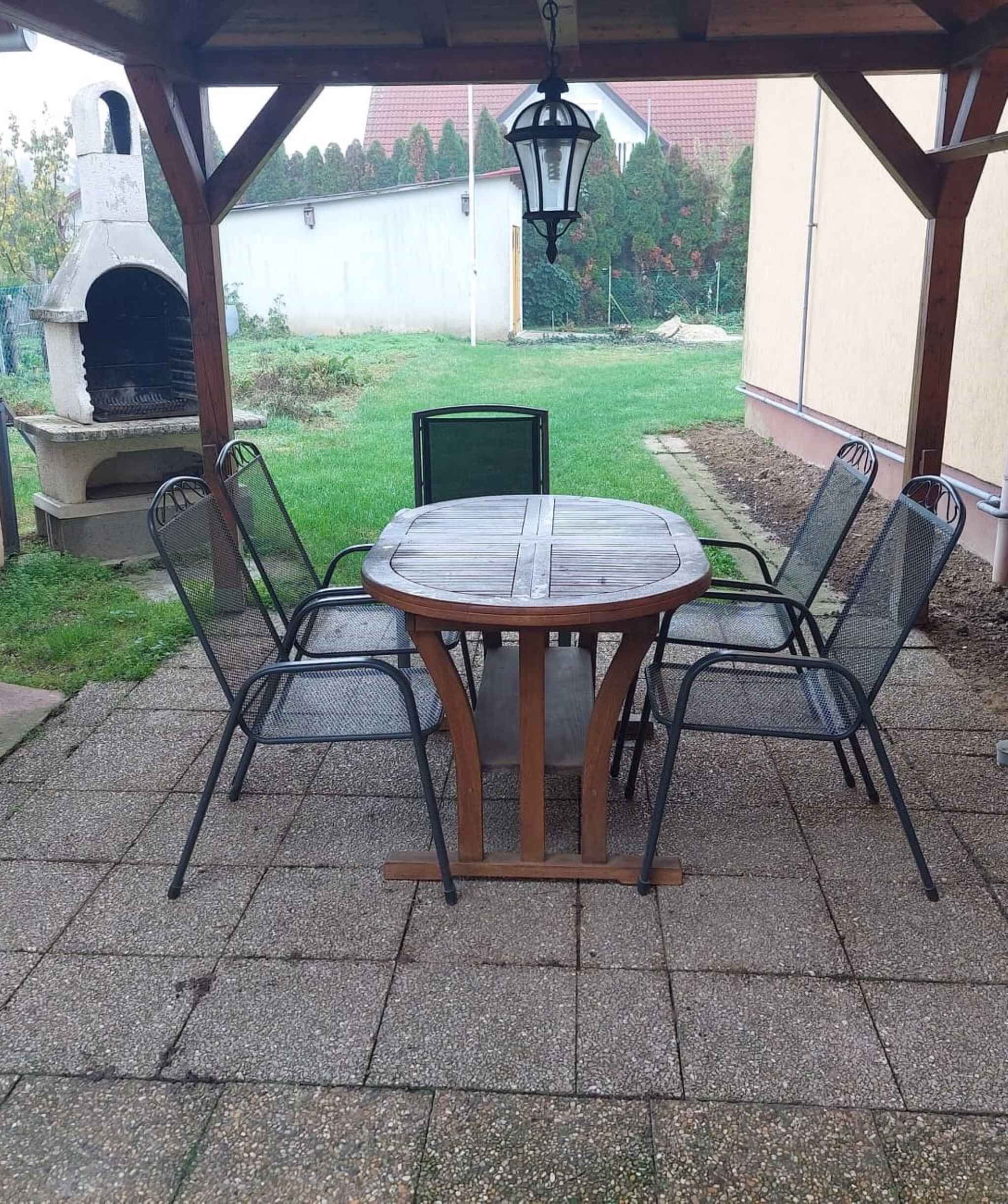 En udendørs terrasse med et rundt bord og fire stole, en hængende lygte og en stenovn.