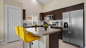 Una cocina moderna con alacenas marrón oscuro, un refrigerador de acero inoxidable, un microondas y un taburete con sillas amarillas.