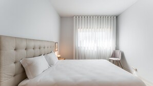 A modern hotel room with a large bed, a nightstand, a chair, and sheer curtains. - GuestReady - Modern apt near Matosinhos Beach (Matosinhos)
