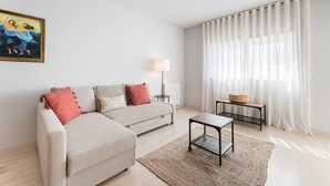 A modern living room with a beige sectional sofa, a small wooden side table, a floor lamp, and a large window with sheer curtains. - GuestReady - Modern apt near Matosinhos Beach (Matosinhos)