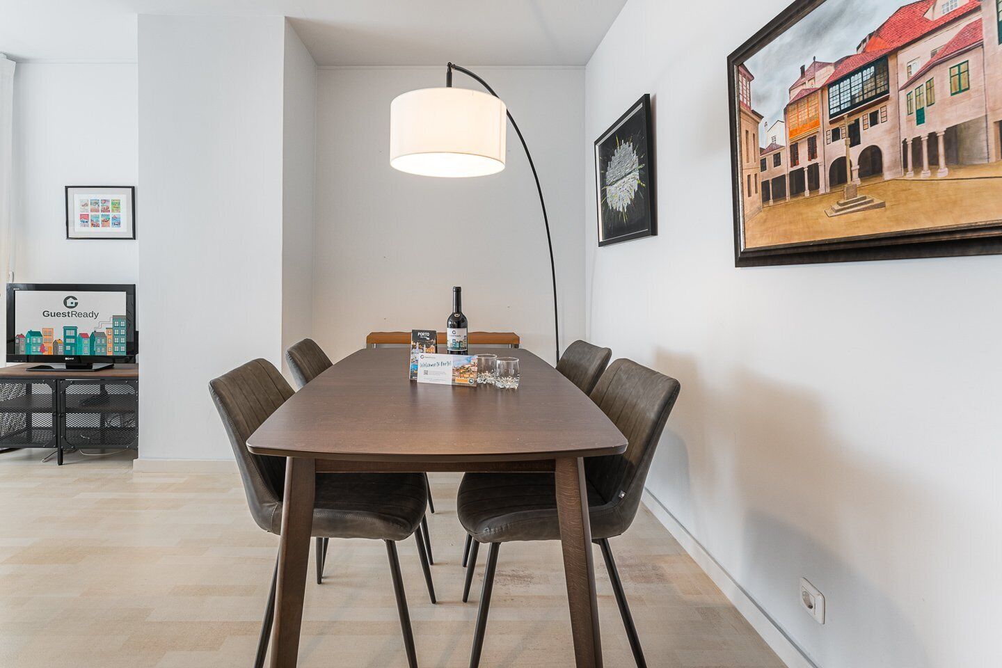 Un comedor moderno con una mesa de madera, seis sillas, una lámpara colgante, un cuadro enmarcado con una vista urbana, y un televisor mostrando 'GuestReady'.