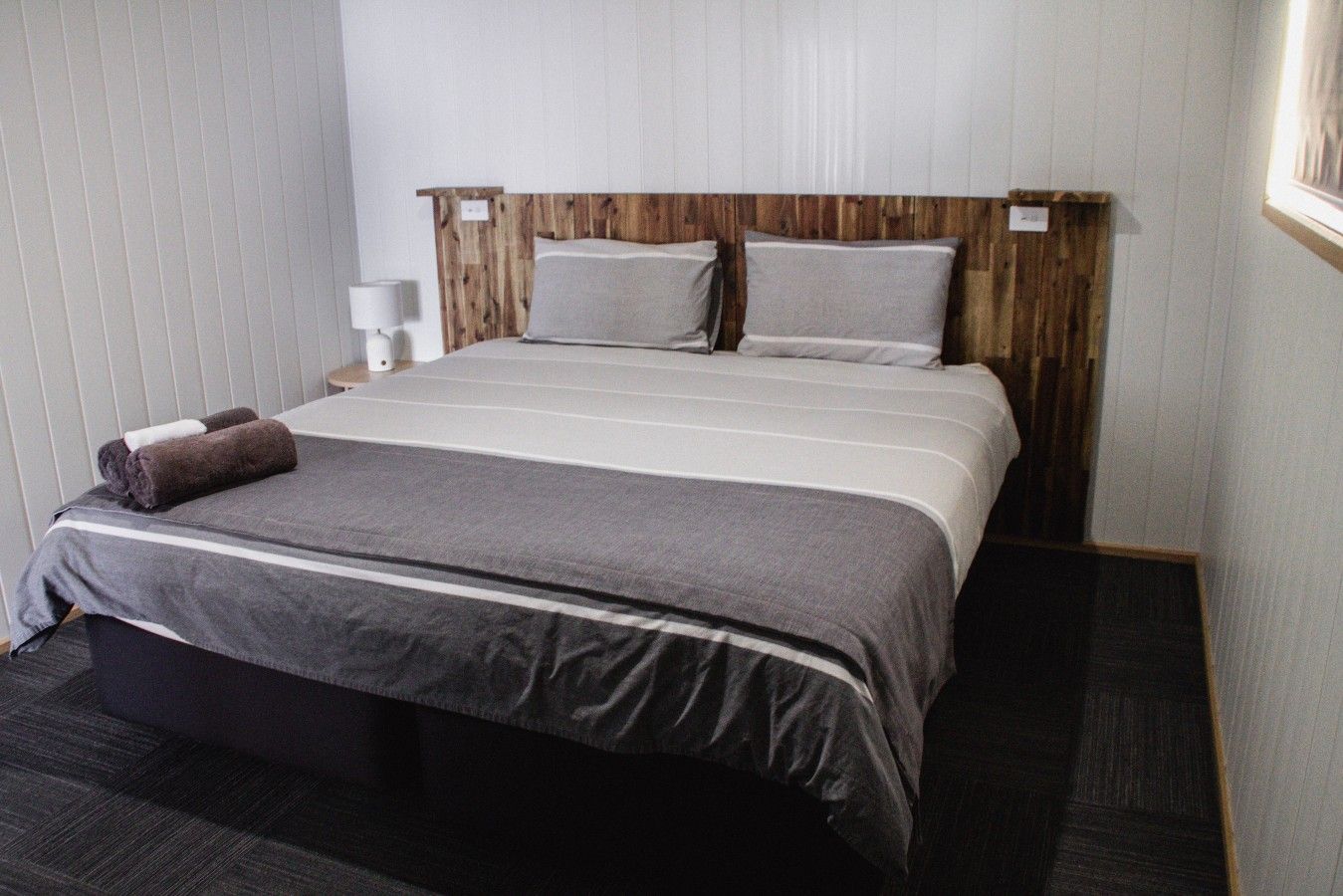 A hotel room with a large bed, a wooden headboard, a bedside table with a lamp, and a dark carpeted floor.