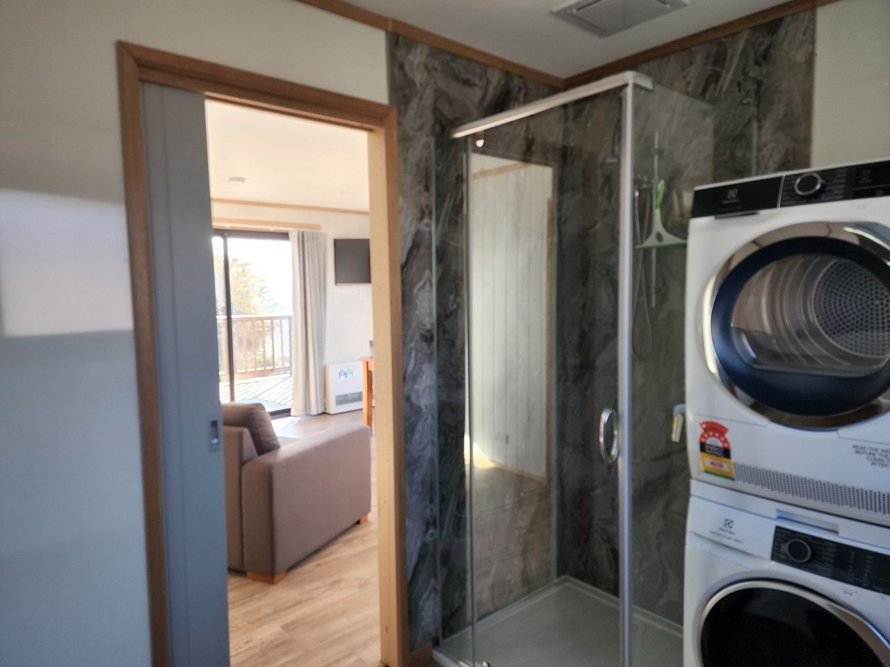 Un baño moderno con cabina de ducha de vidrio, lavadora integrada y vista a un área de estar a través de una puerta abierta.