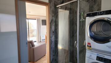 Un baño moderno con cabina de ducha de vidrio, lavadora integrada y vista a un área de estar a través de una puerta abierta.