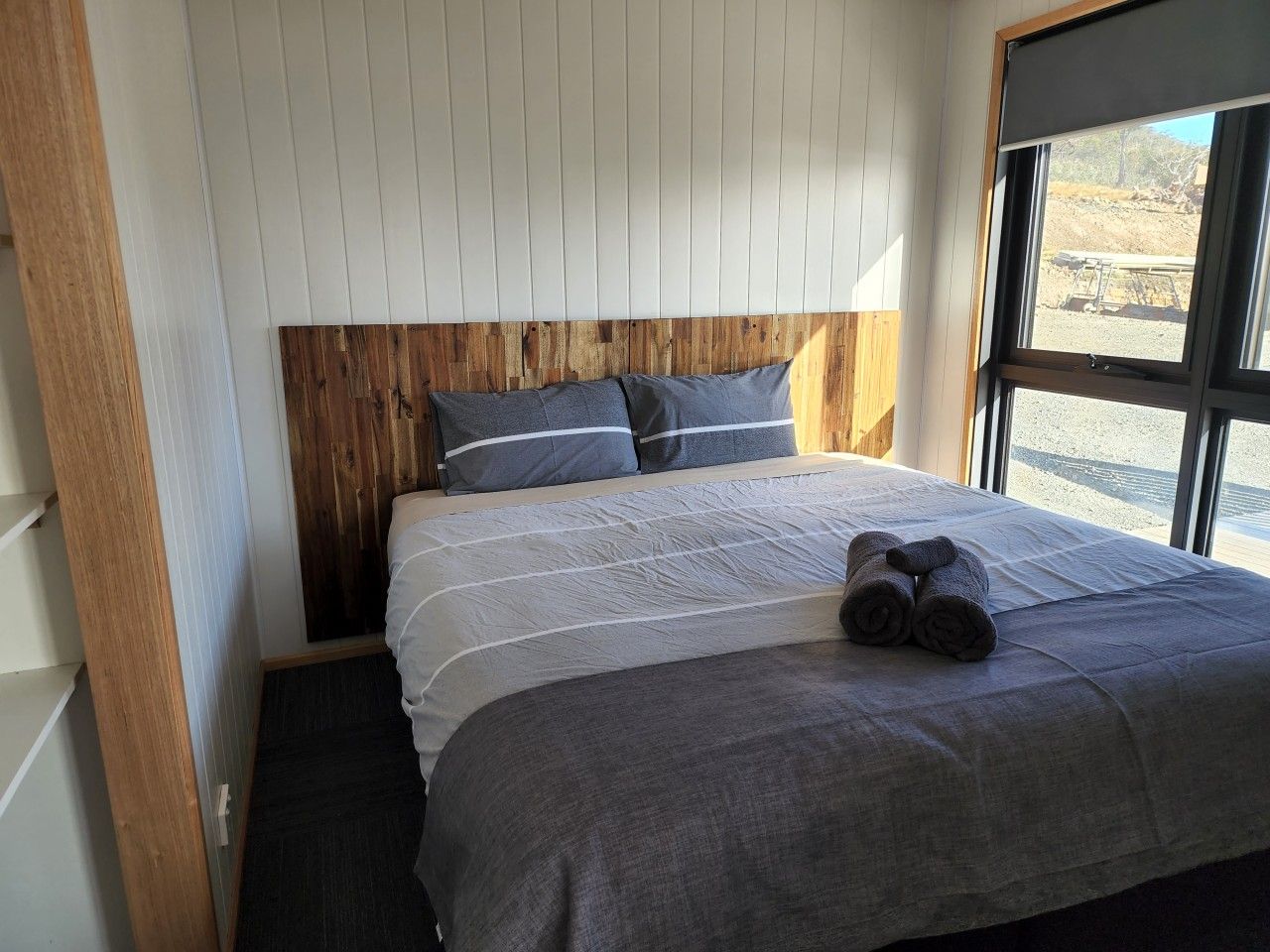 A bedroom with a wooden headboard, a bed with grey bedding, and a window with a view of the outdoors.