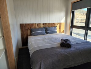 Een slaapkamer met een houten hoofdbord, een bed met grijze beddengoed en een raam met uitzicht op het landschap.