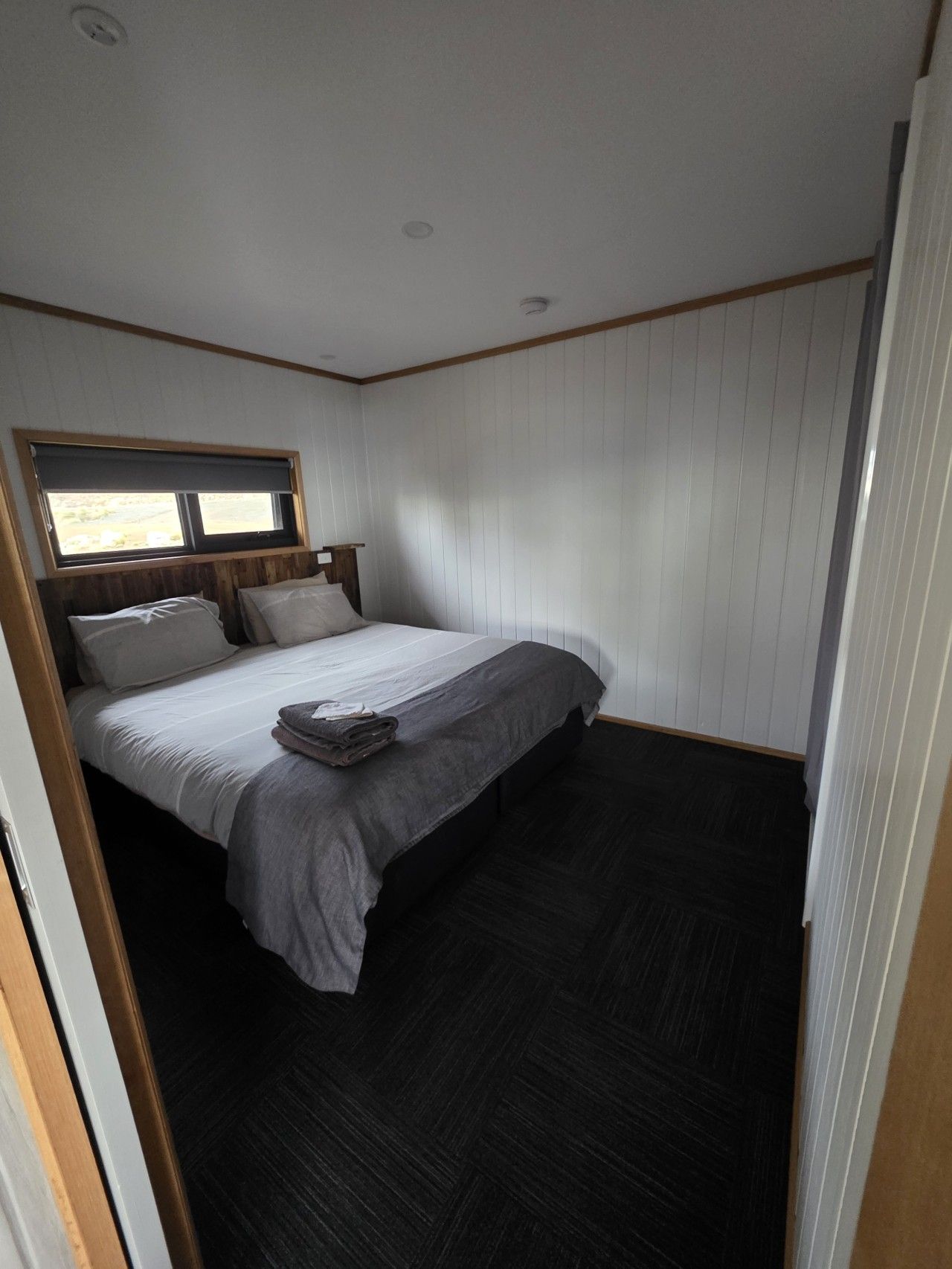 Ein Einzelbett mit Holz-Kopfstück, ein Fenster mit Jalousien und ein dunkler Boden.