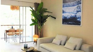 Un salon moderne avec un canapé, une table basse et une grande plante. Un tableau orne le mur, et on aperçoit un coin repas au fond de la pièce.